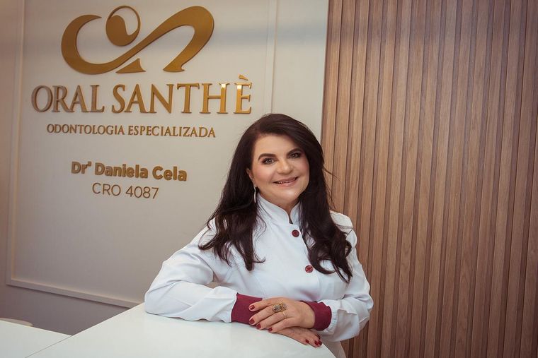 Dra. Daniela Cela de jaleco, diante da parede com a marca Oral Santhè